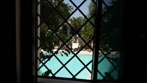Vista a través de una ventana con una celosía de diamantes, que da a una piscina turquesa y un edificio de piedra con una cerca blanca.