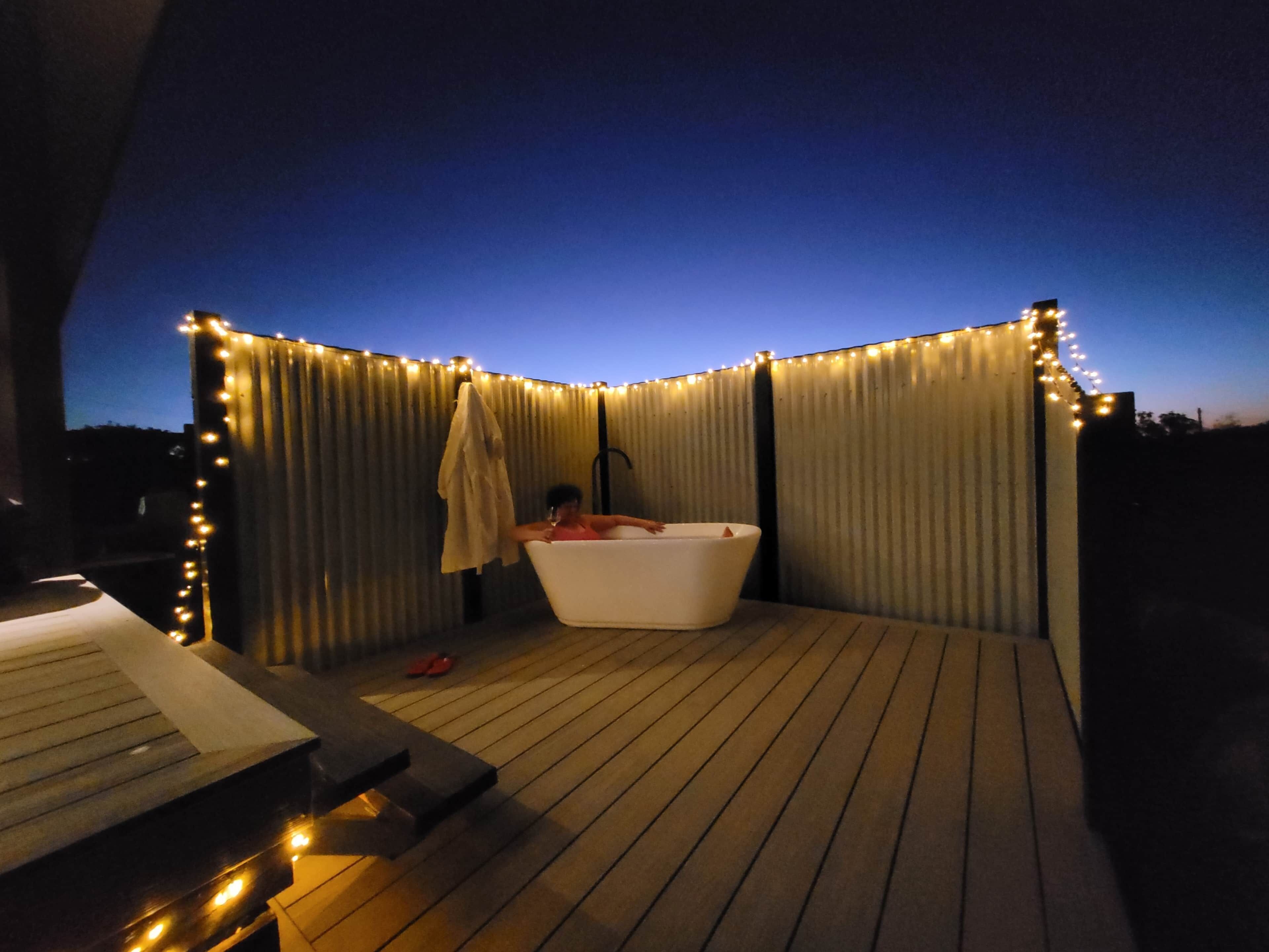Une terrasse en bois avec une baignoire blanche, un fauteuil de relaxation et des guirlandes lumineuses.
