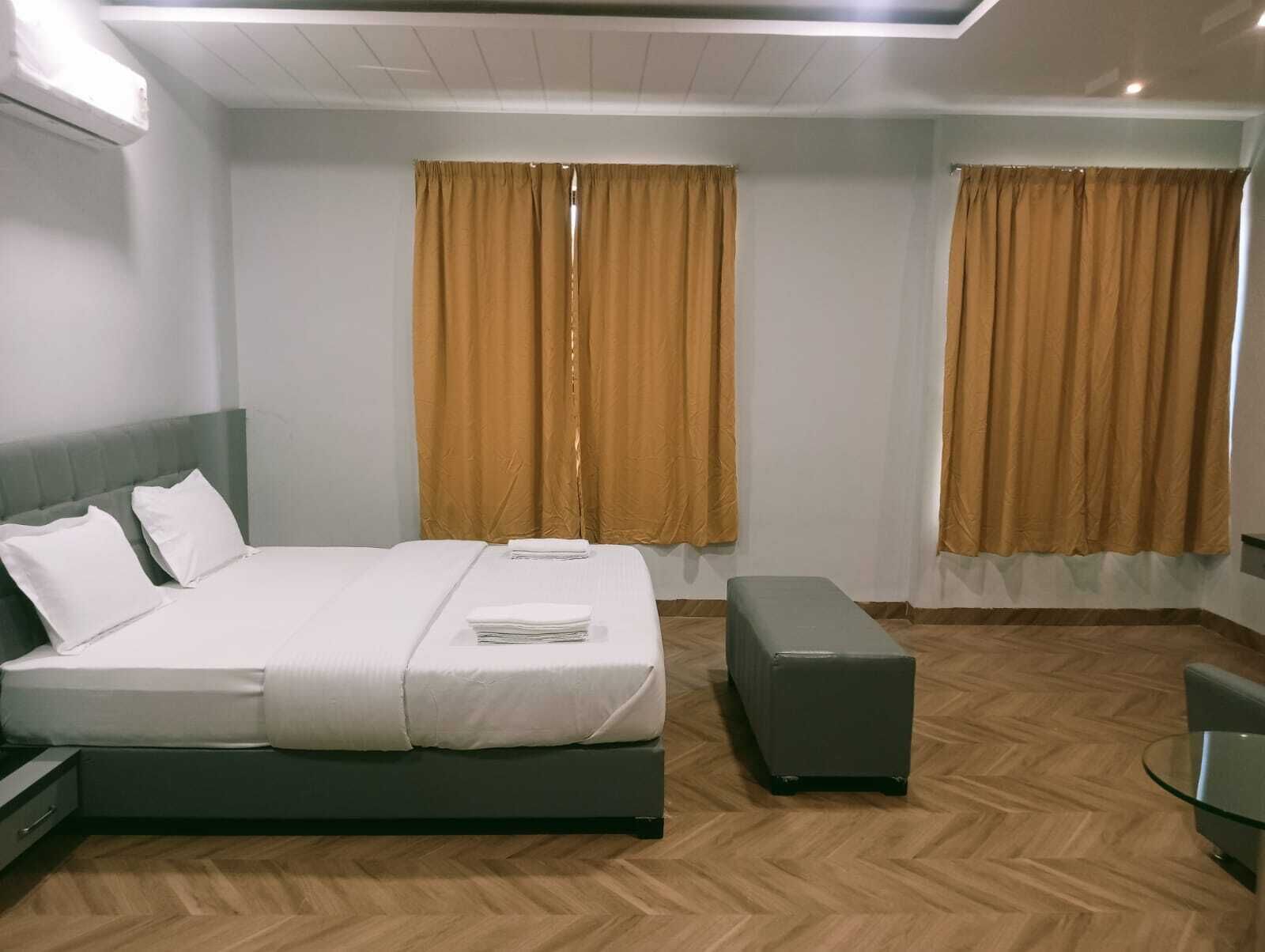 A hotel room with a bed, a bench, a small table, and two windows with curtains.
