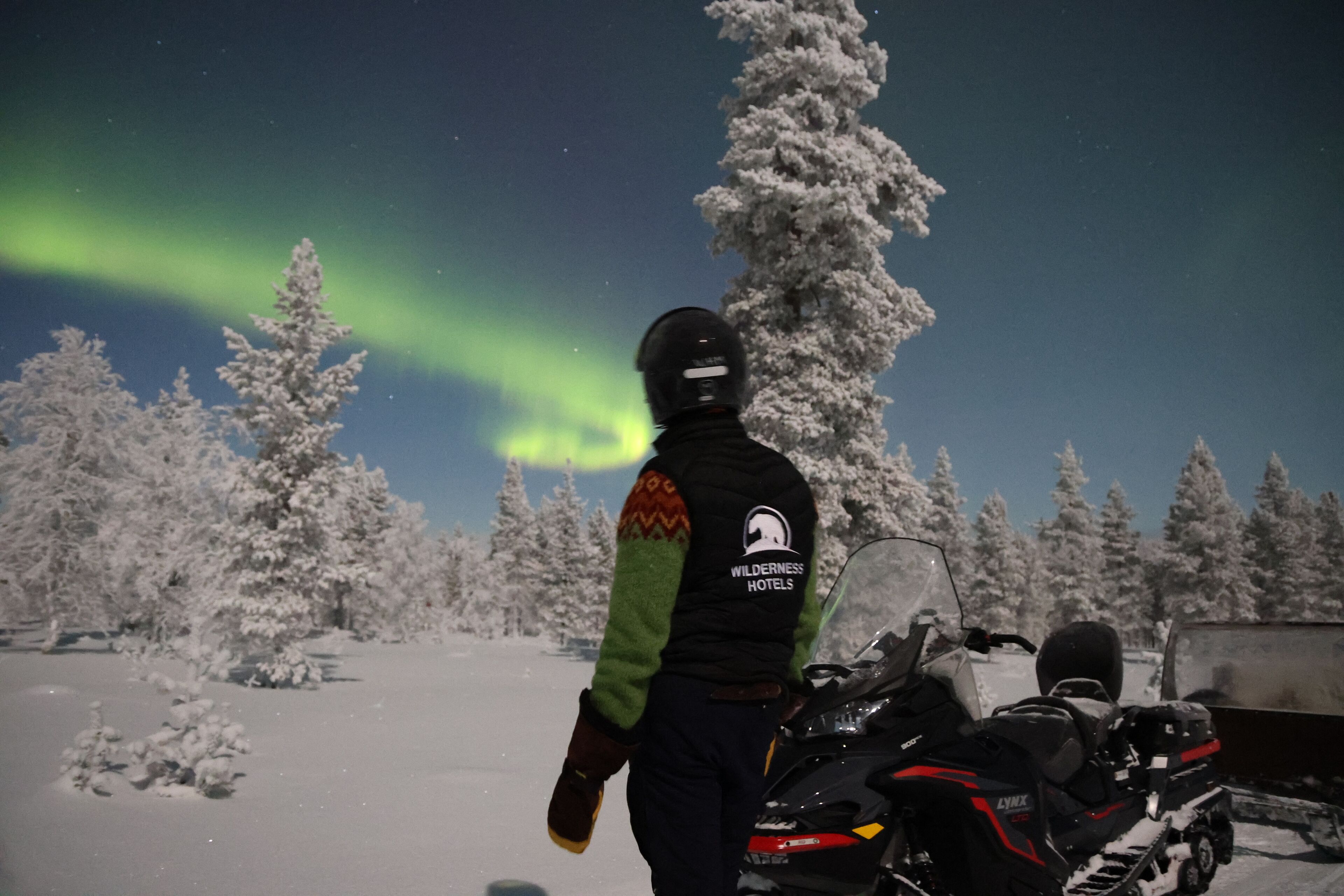 A person standing next to a snowmobile in a snowy forest with trees and a clear night sky.