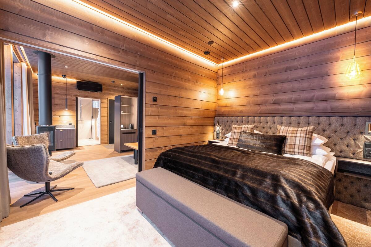 Ein modernes Schlafzimmer mit Holzboden, einem großen Bett mit einer gepolsterten Kopfteil sowie einer Sitzecke mit einem Sessel und Blick ins Badezimmer.