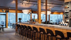 Een barruimte met houten balken, barstoelen en uitzicht op besneeuwde bomen door grote ramen.