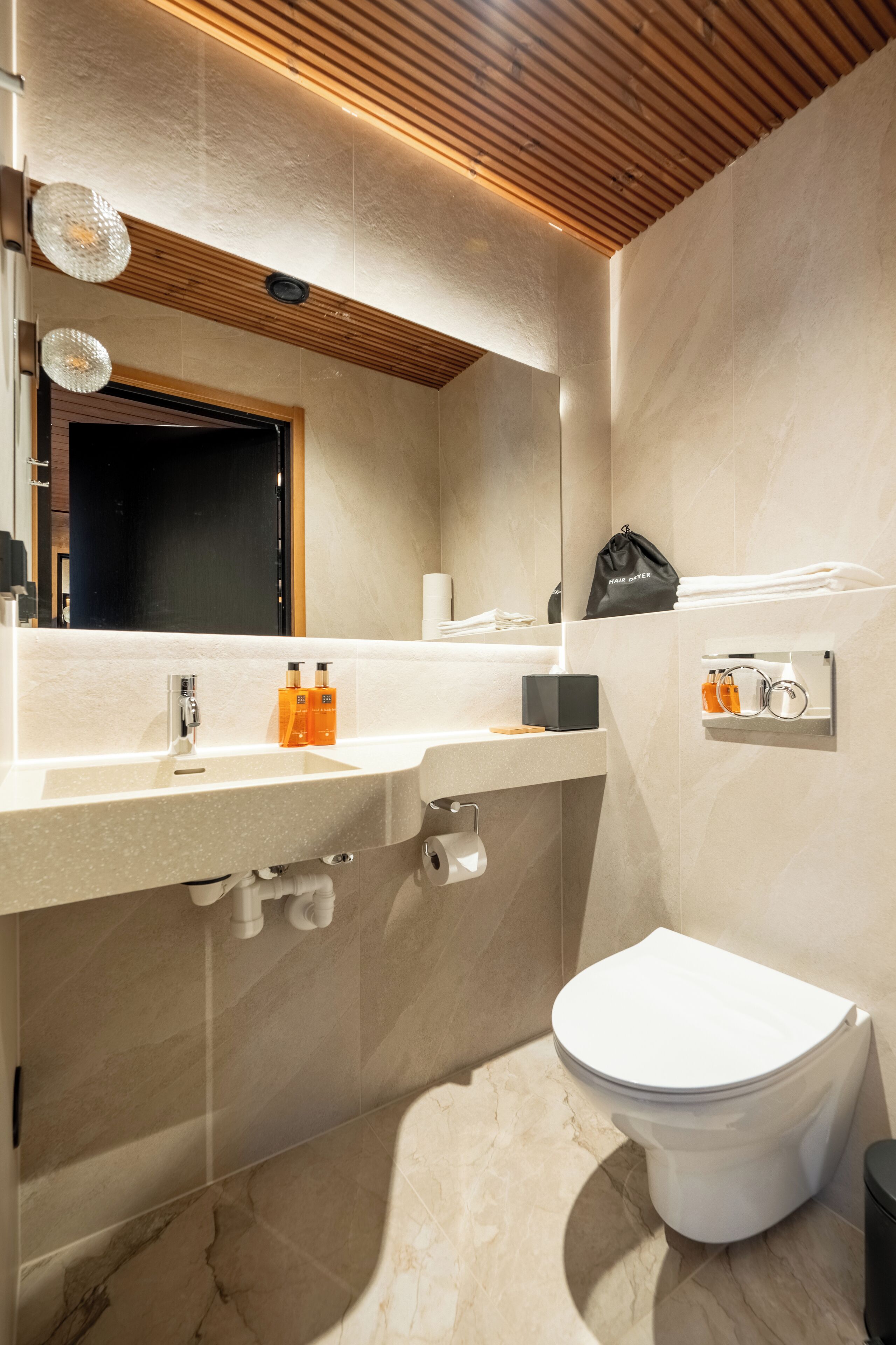 A modern bathroom with a wooden ceiling, a large mirror, a toilet, and a sink with toiletries.