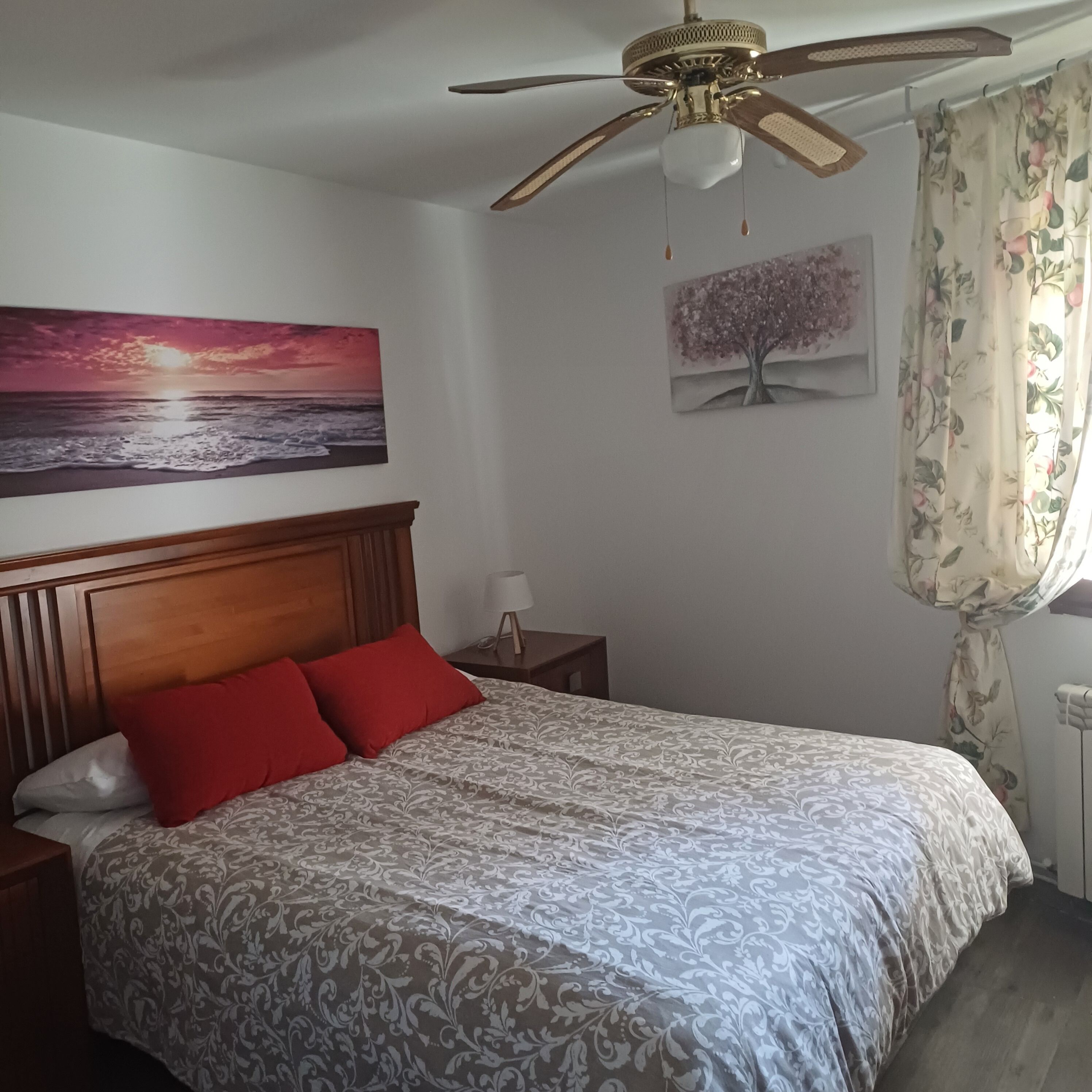 A bedroom with a bed, a ceiling fan, a nightstand, a lamp, a painting of a sunset, and a floral curtain.