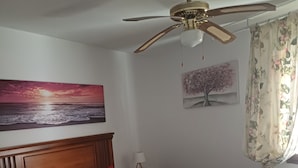 Camera da letto con un letto, una ventola a soffitto, un comodino, una lampada, un quadro raffigurante un tramonto e una tenda floreale.