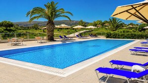 A swimming pool with blue water, surrounded by lounge chairs and umbrellas, with a palm tree and hills in the background. - Nautilos Hotel (Kefalonia)