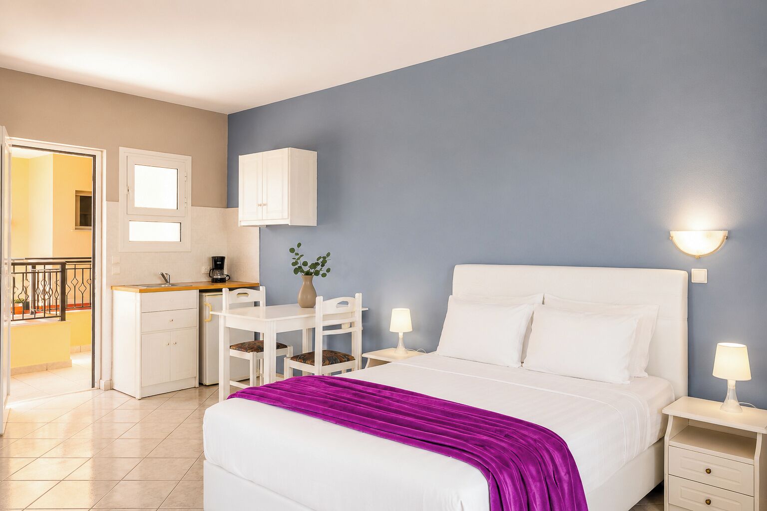 Een hotelkamer met een bed, nachtkastjes, een bureau met stoel, een kleine plant en uitzicht op de keuken.
