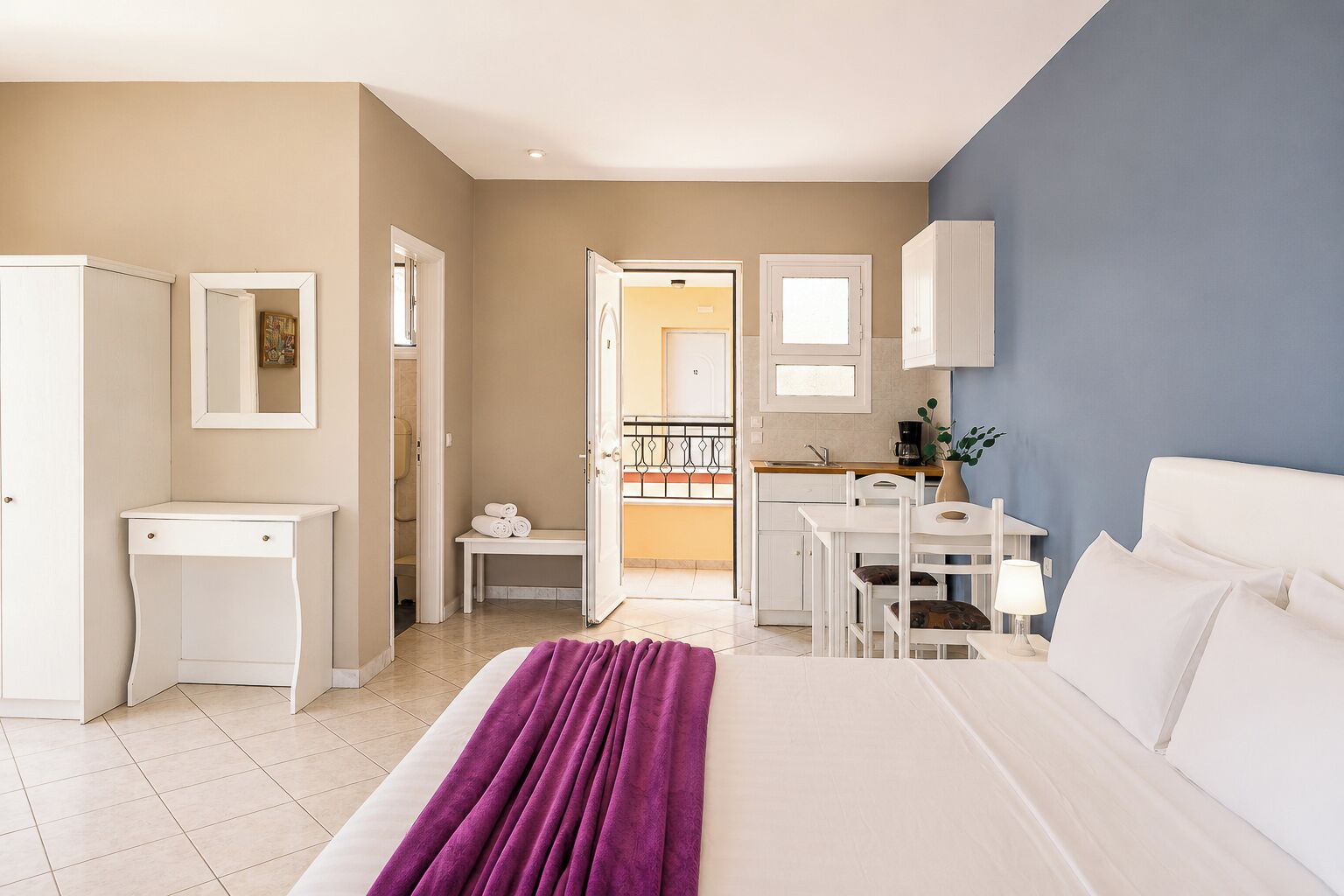 A hotel room with a bed, a desk, a chair, a small table, and a bathroom visible through an open door.