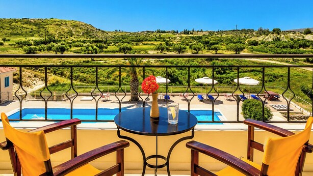 A balcony with a view of a pool and landscape, a table with chairs, and a vase with flowers. - Nautilos Hotel (Kefalonia)