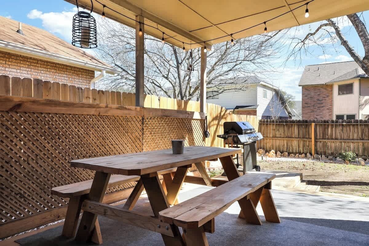 Une terrasse extérieure avec une table de pique-nique et des bancs en bois, un barbecue et un espace abrité agrémenté de guirlandes lumineuses.