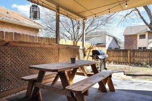 An outdoor patio with a wooden picnic table and benches, a barbecue grill, and a covered area with string lights. - Cozy 3/2 Stylish home great for families (San Antonio)