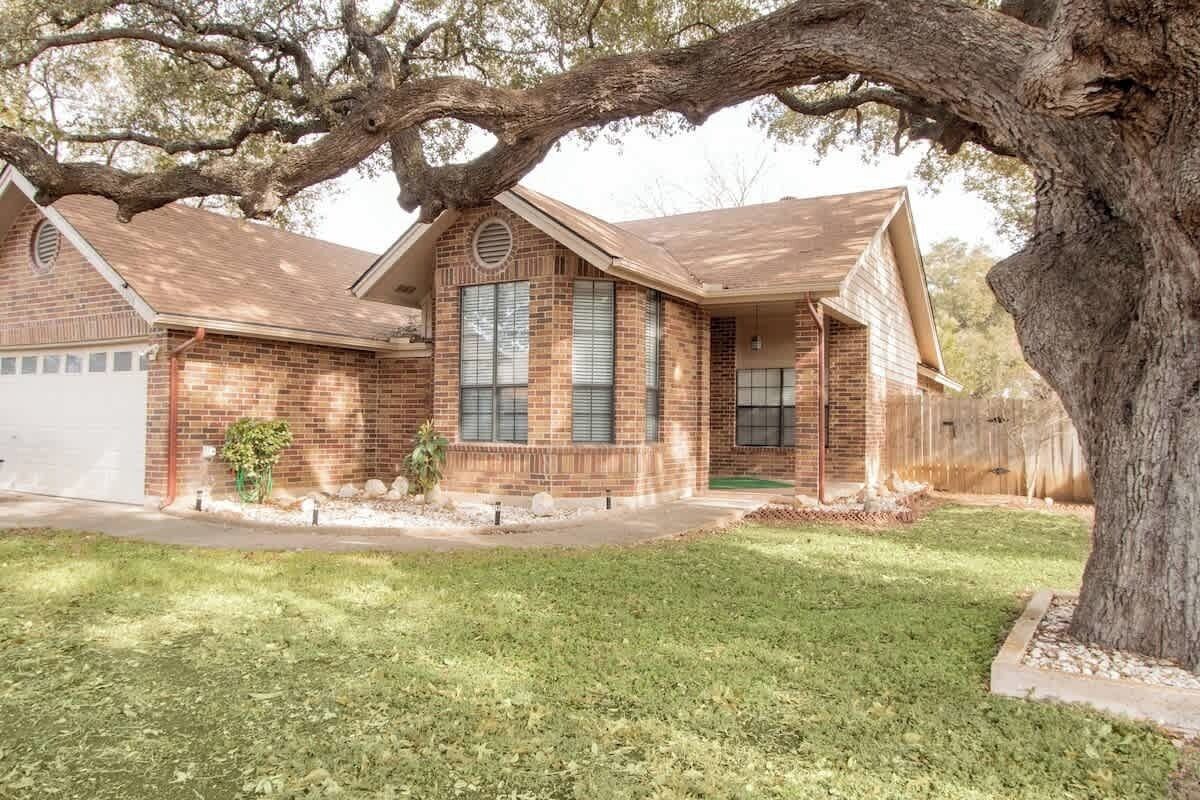 A single-story brick house with a front porch, a garage, and a large tree in the front yard.