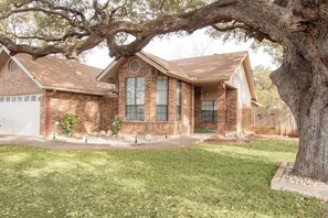 A single-story brick house with a front porch, a garage, and a large tree in the front yard. - Cozy 3/2 Stylish home great for families (San Antonio)