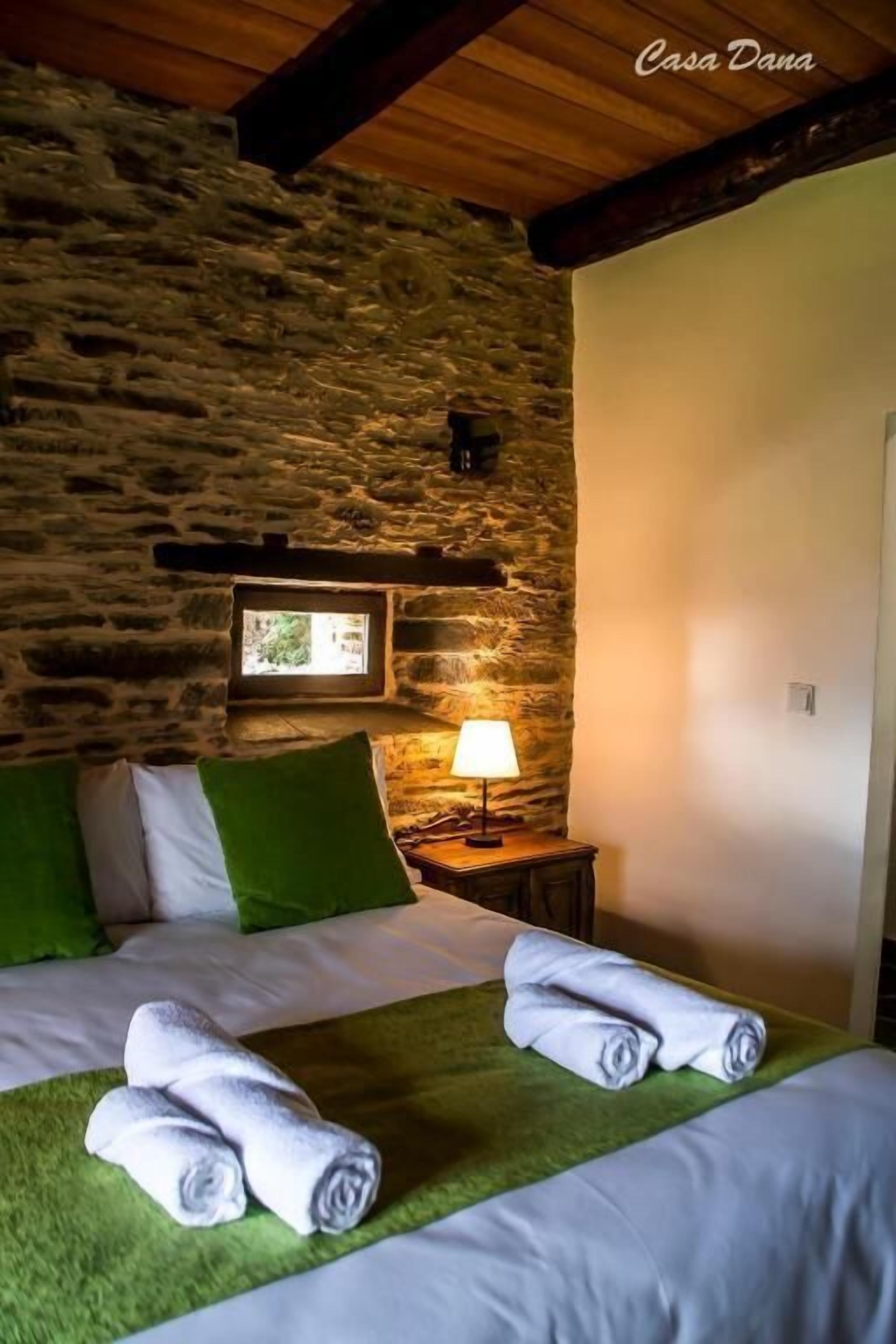 A bedroom with a stone wall, a bed with green and white bedding, a lamp, and a window.