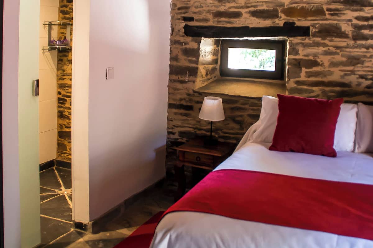 A bedroom with a stone wall, a bed with white and red bedding, a nightstand with a lamp, and a window with a view.