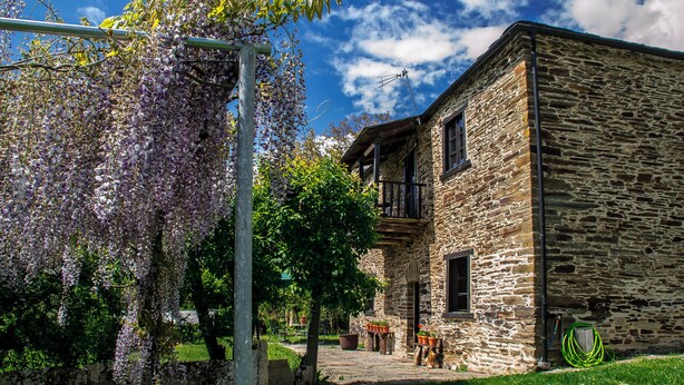 Um edifício de pedra com varanda, cercado por vegetação e trepadeiras de glicínia.