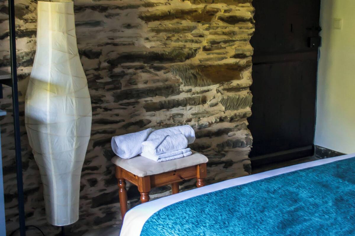 A room with a stone wall, a bed with a blue cover, a bedside table with a towel, and a door.