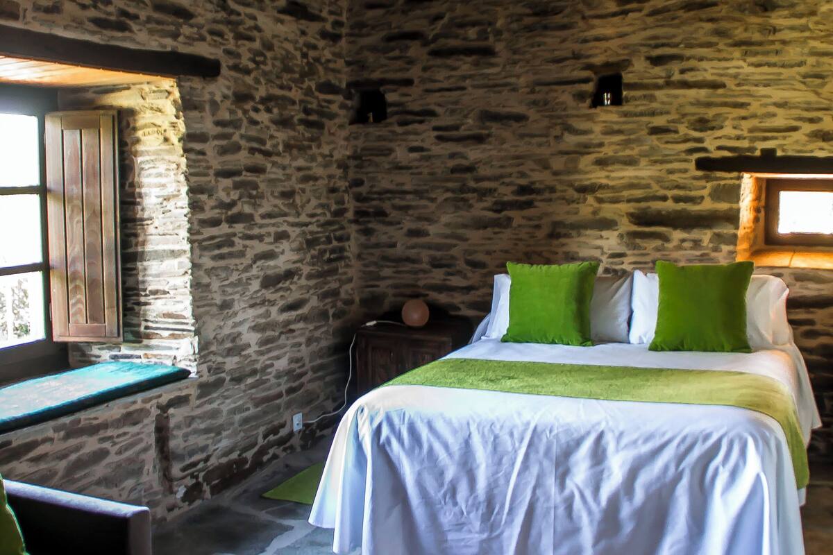 A rustic room with a stone wall, a wooden ceiling, a bed with green pillows, and a small table with a tray.