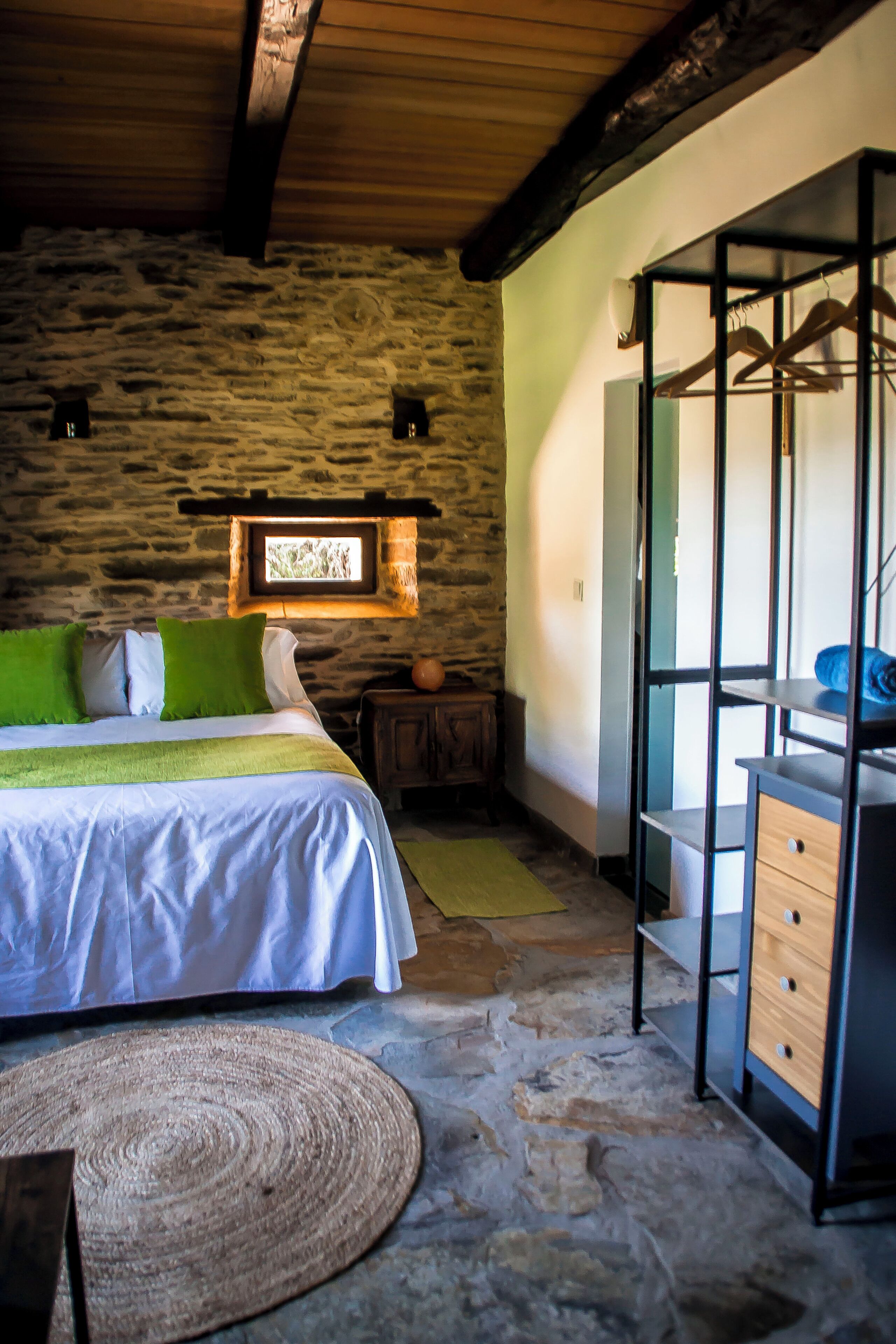Une chambre avec un mur en pierre, un lit avec une literie verte et blanche, une commode en bois et un luminaire suspendu.