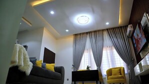 A modern living room with a sofa, a yellow armchair, a coffee table, and a TV. - Lush Meadows Apartments (Abuja)