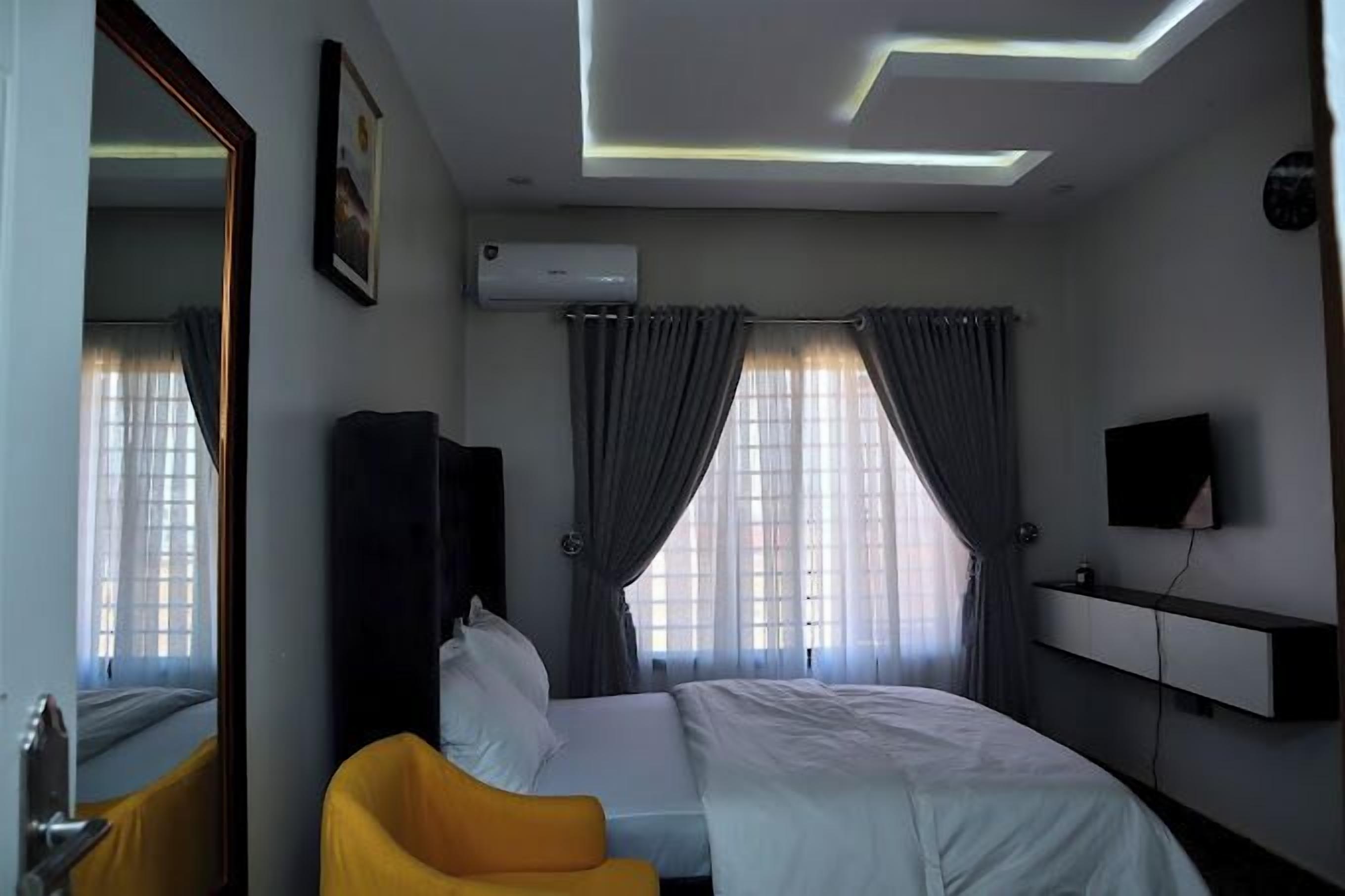 A hotel room with a bed, a yellow armchair, a flat-screen TV, and a ceiling with recessed lighting.
