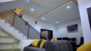 A modern living room with a staircase, a sofa with cushions, a dining table with chairs, and a clock on the wall. - Lush Meadows Apartments (Abuja)
