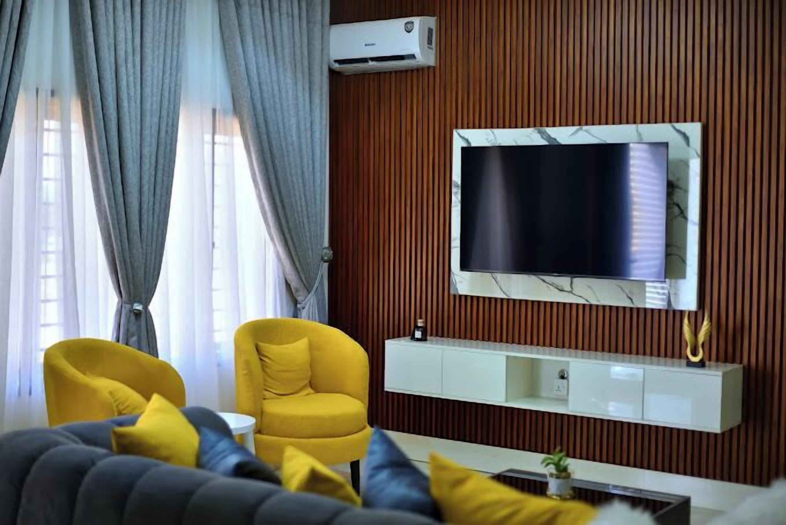 A modern living room with a large flat-screen TV mounted on a wooden wall, a sofa with yellow and grey cushions, and a yellow armchair.