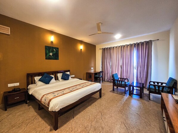 Habitación de hotel con cama, escritorio, dos sillas, ventilador de techo y ventana con cortinas.