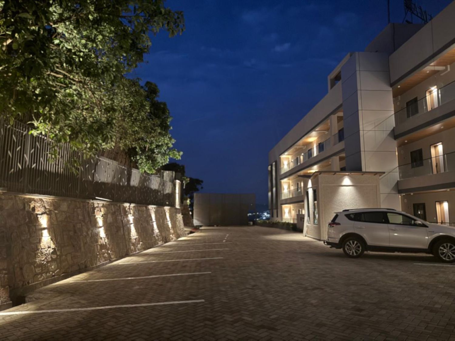 A modern building with a car parked in front, a paved pathway, and a stone wall on the left.