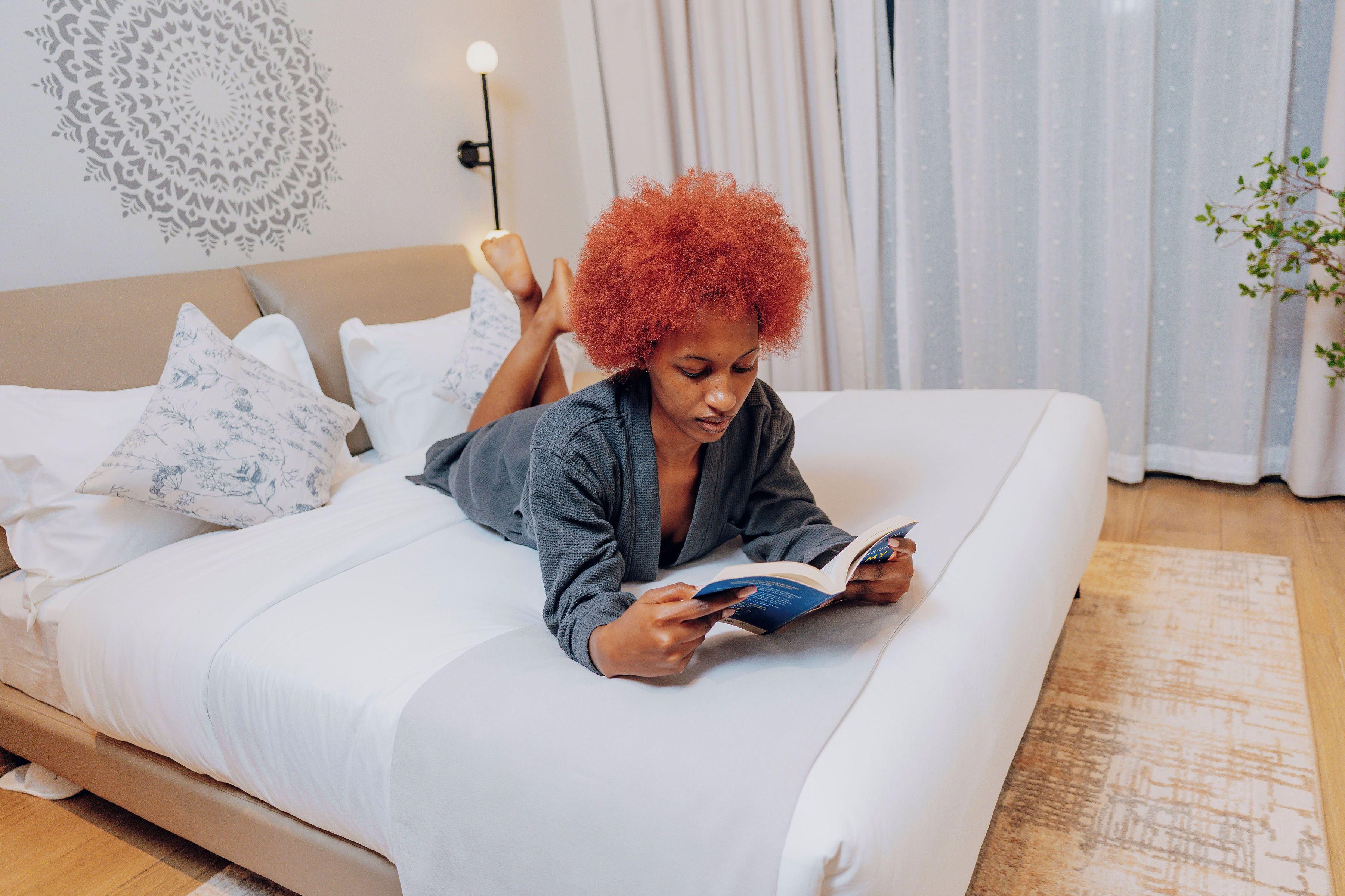 Una persona acostada en una cama, un libro, un almohadón con estampado floral y una pared con diseño circular.