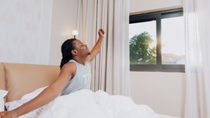 Une personne est allongée dans un lit recouvert d’une couette blanche. Une fenêtre avec des rideaux est visible, ainsi qu’un mur orné d’un motif de feuilles.