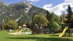 Une aire de jeux avec des toboggans et des structures d’escalade, entourée d’une pelouse et d’arbres, avec des montagnes en arrière-plan.