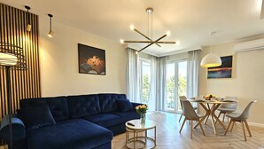 A modern living room with a dark blue sofa, a round coffee table, and a dining area with a table and chairs. There is a painting on the wall, a ceiling light fixture, and an air conditioning unit. - Apartament Luxe w Kołobrzegu - 365PAM (Kolobrzeg)