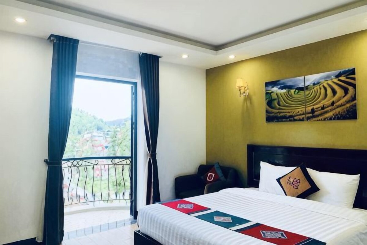 A hotel room with a bed, a view of the outdoors, a painting on the wall, and a balcony with curtains.