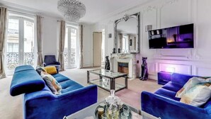 A spacious living room with two blue sofas, a glass coffee table, a chandelier, a fireplace, and a large mirror. - AN AUTHENTIC PARISIAN ELEGANCE MOMENTS FROM ICONIC LANDMARKS – CHAMPS ELYSEES (Paris)