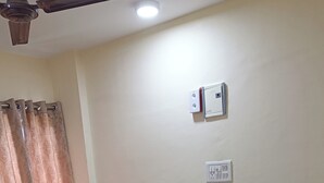 A bedroom with a bed, two pillows, a nightstand, a power outlet, a ceiling fan, and a window with curtains. - Divine Residency (Panvel)