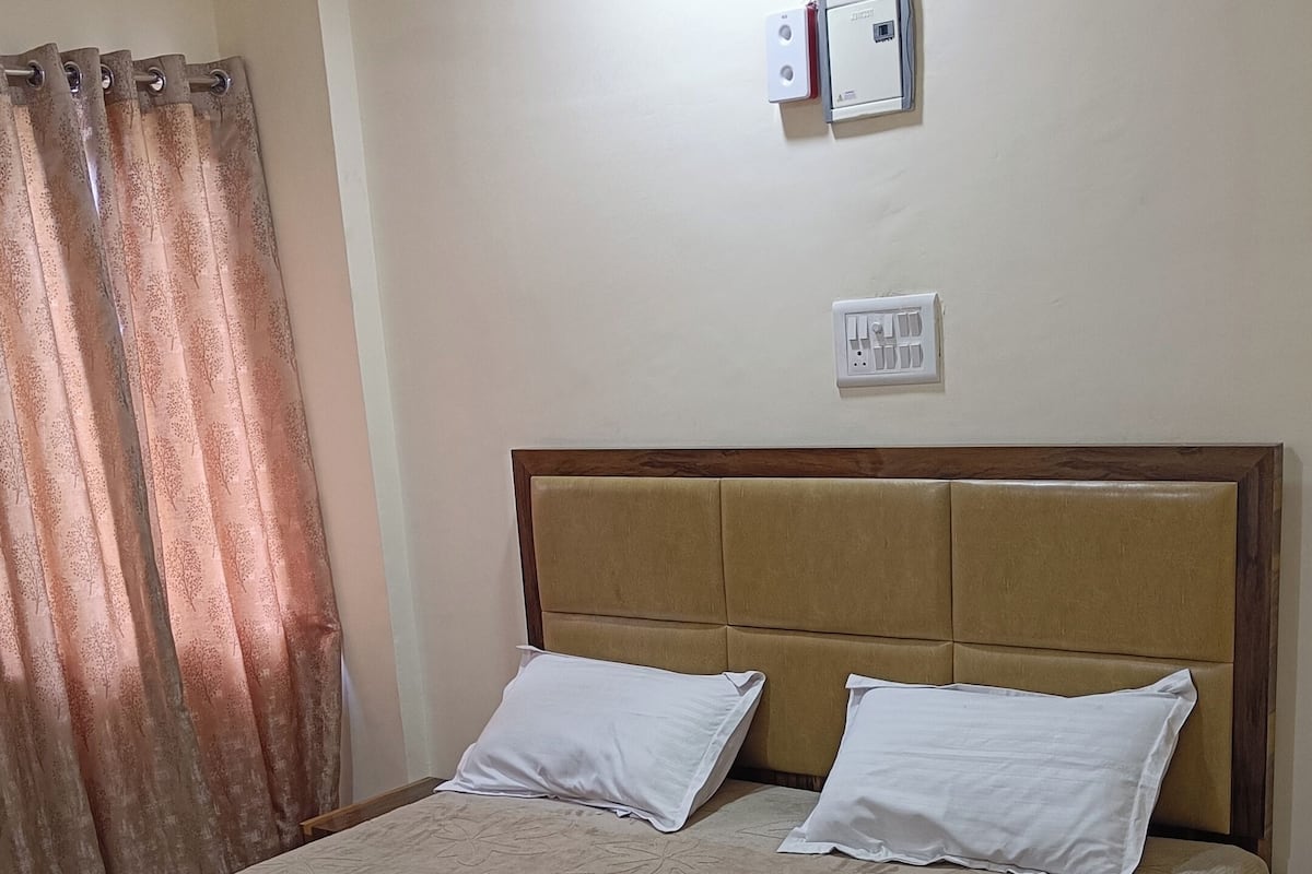 A bedroom with a bed, two pillows, a nightstand, a power outlet, a ceiling fan, and a window with curtains.