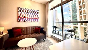 Une chambre d’hôtel moderne dotée d’une grande fenêtre donnant sur la ville, d’un canapé marron avec des coussins rouges, d’une table ronde blanche et d’un tableau abstrait aux couleurs vives accroché au mur.