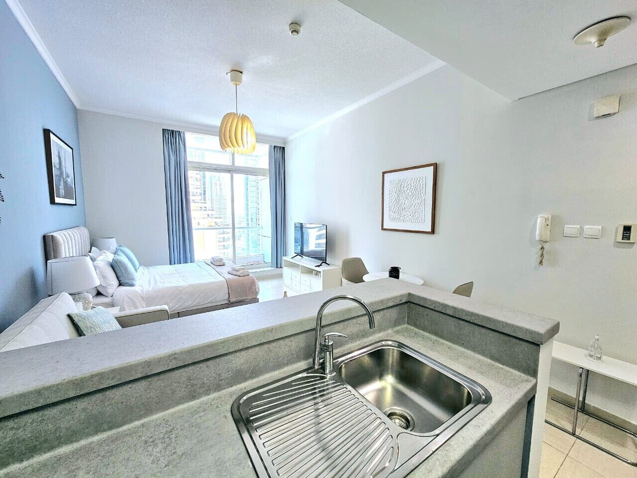 Una habitación de hotel moderna con kitchenette, sofá cama, zona de comedor y un gran ventanal con cortinas.