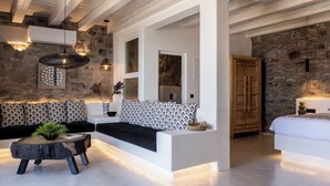 A modern interior with a stone wall, a white sofa, a round wooden table with a plant, and a bed with a white headboard. - Makani Suites (Mykonos)