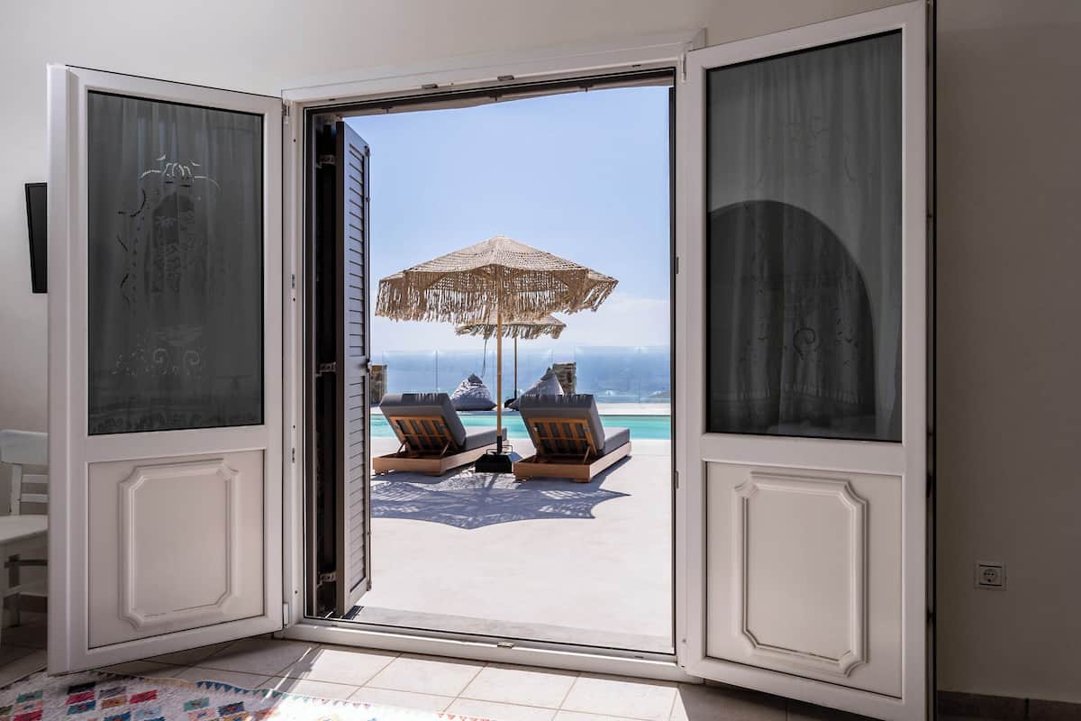 An open door leading to a balcony with two lounge chairs and a straw umbrella, overlooking a clear blue sea.