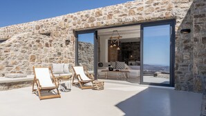 A stone exterior with a glass door leading to a modern interior with a sofa and a dining table. - Makani Suites (Mykonos)