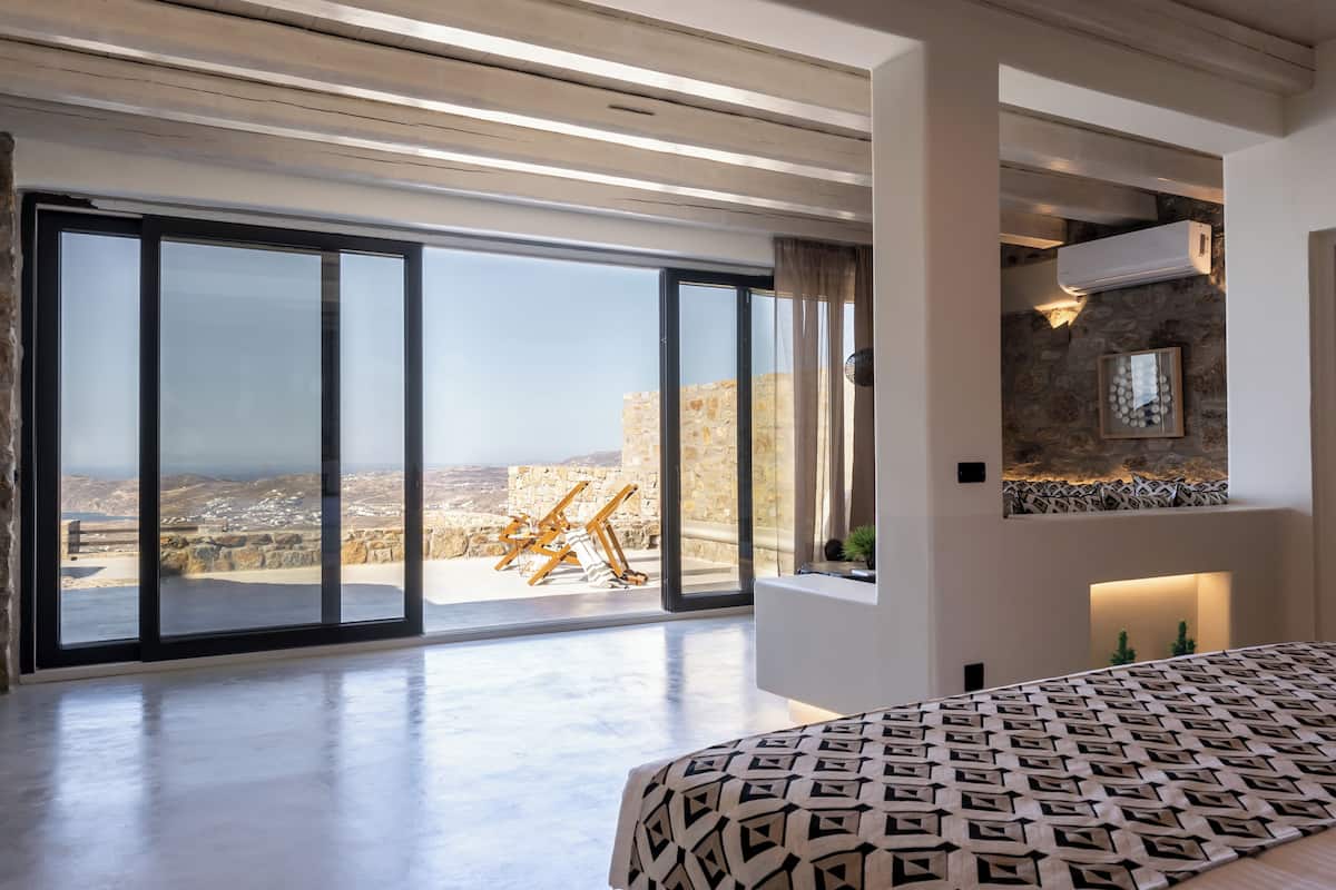 A spacious bedroom with a large bed, a view through floor-to-ceiling windows, and a stone wall with a decorative light fixture.