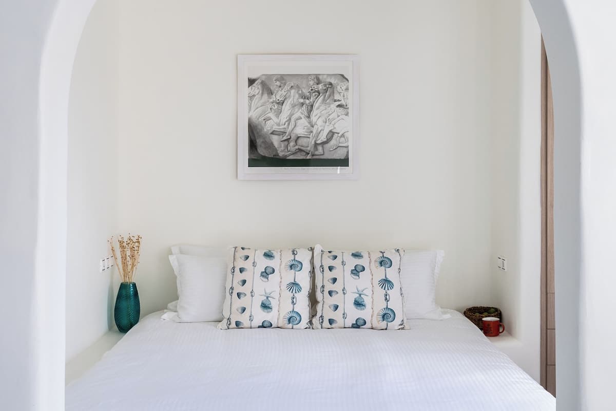 A bedroom with a white bed, patterned pillows, a framed artwork, a vase with dried plants, and a basket.