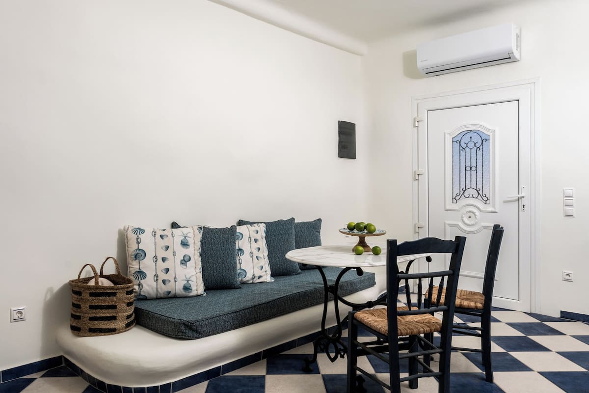 A room with a sofa, a small table with a fruit bowl, a chair, a wall-mounted air conditioner, and a door with a decorative design.