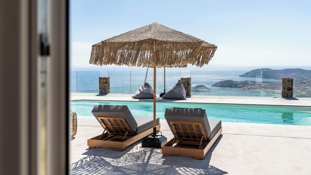 A poolside area with two lounge chairs and a straw umbrella, overlooking a clear blue pool and distant rocky landscape. - Makani Suites (Mykonos)