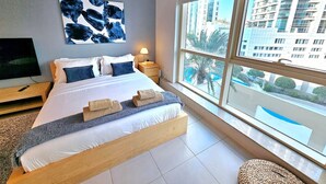 Une chambre d’hôtel moderne avec un grand lit, un téléviseur posé sur un meuble, un tableau au mur et une vue sur la ville grâce à de grandes fenêtres.