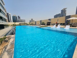 Ein Dachpool mit klarem, blauem Wasser, umgeben von Liegestühlen und Sonnenschirmen, mit modernen Gebäuden im Hintergrund.