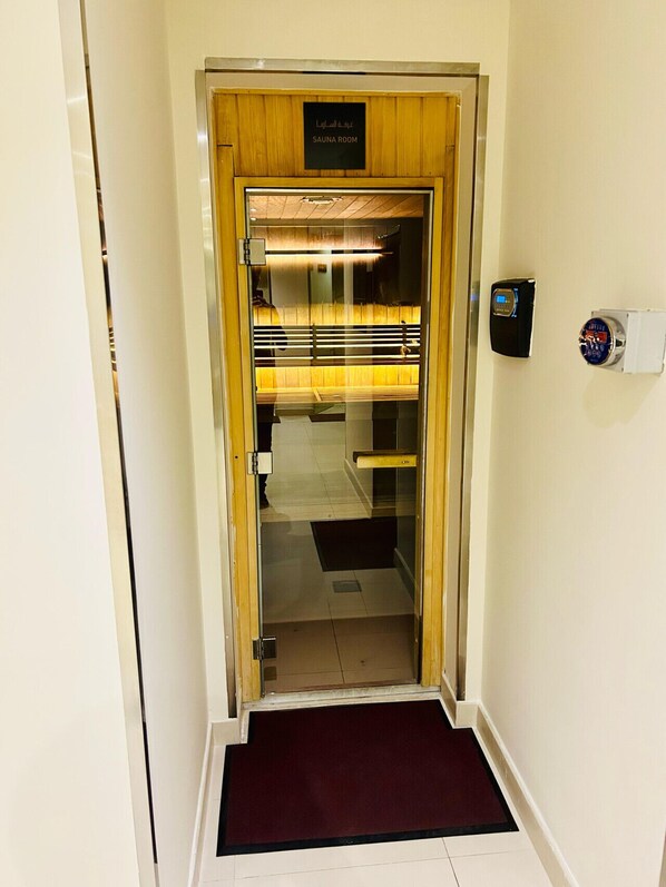 A sauna room entrance with a wooden door, a digital control panel, and a red mat at the bottom.