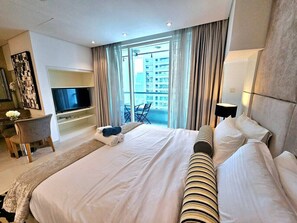 A hotel room with a large bed, a TV, a desk, and a balcony with a view of the city.